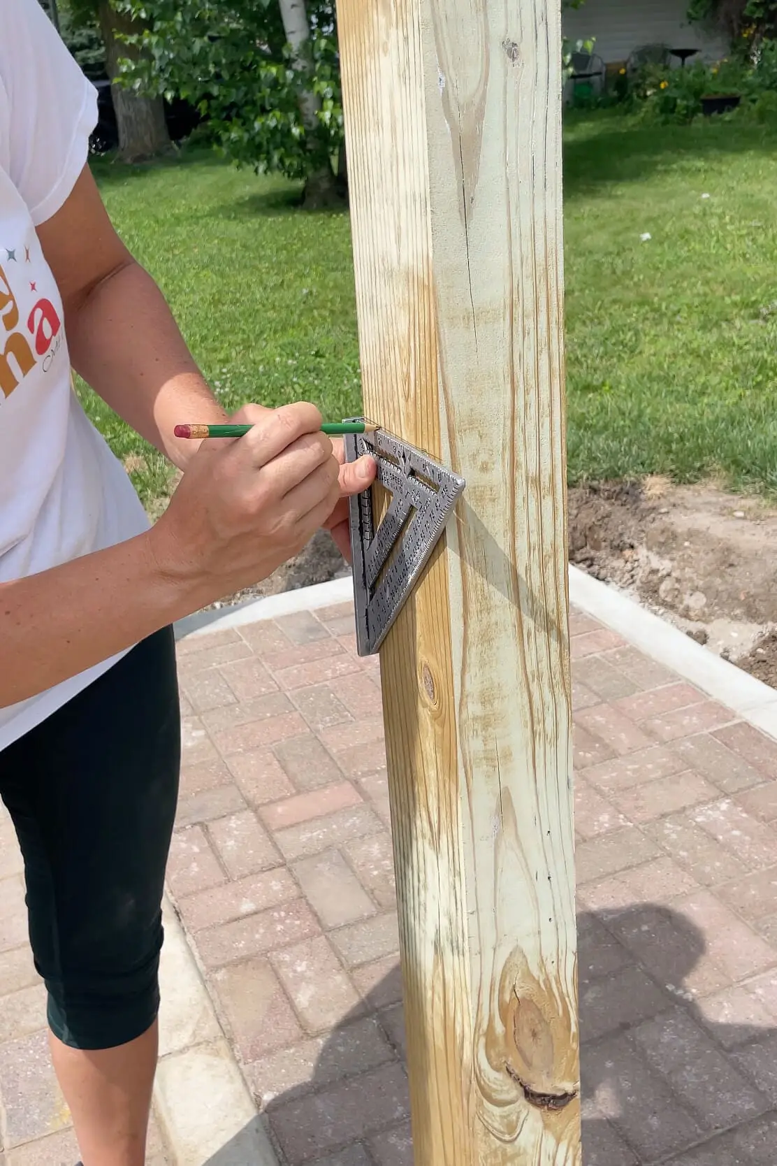 DIYer using a speed square and pencil to mark precise cut lines on a vertical wood post for a trash enclosure privacy fence.